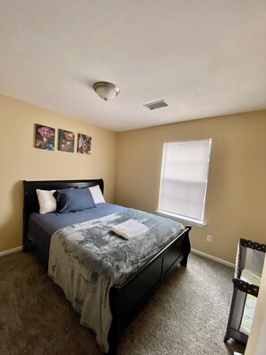 Un dormitorio con cama, una ventana con persianas, una lámpara de techo y tres cuadros colgados en la pared.