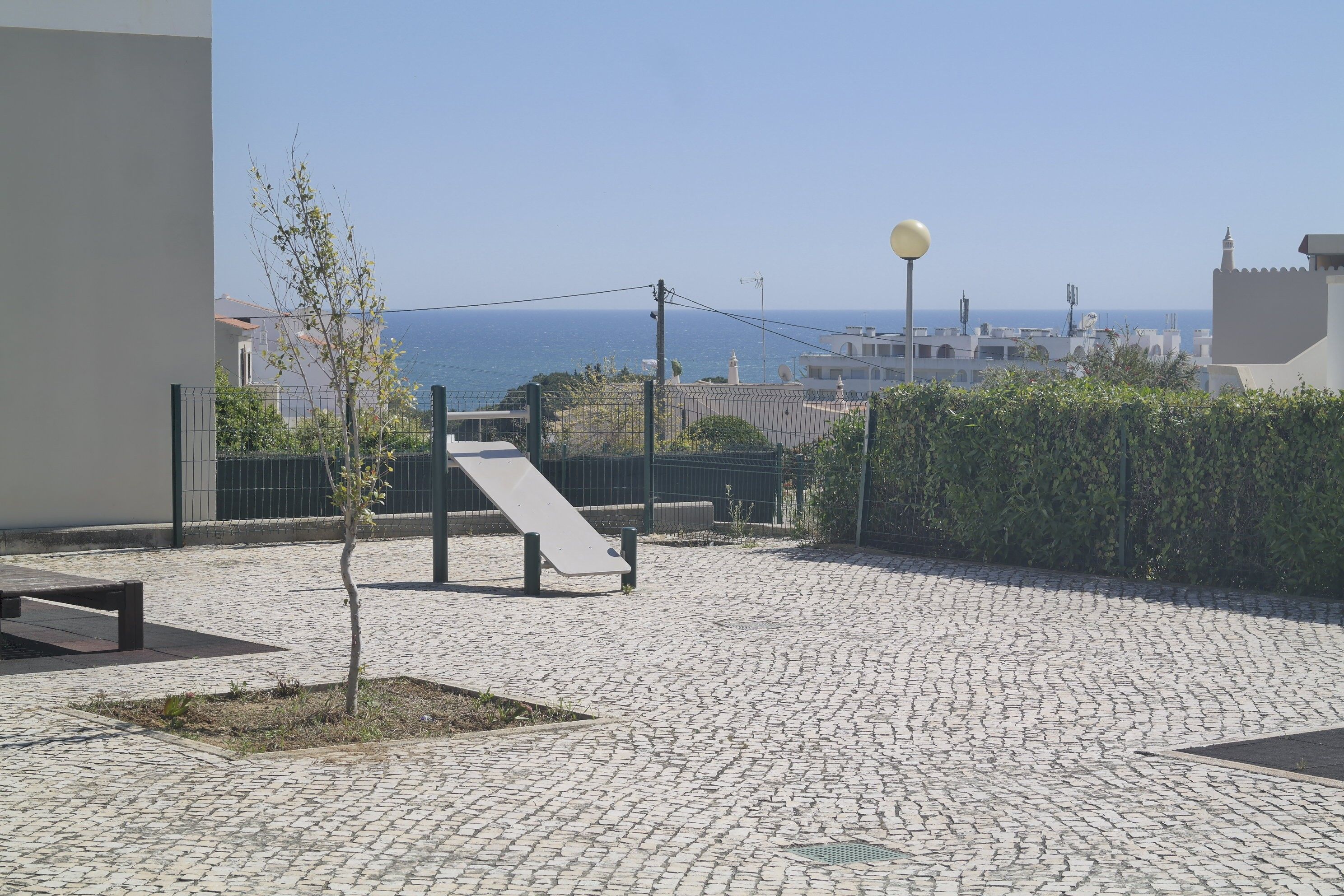 A paved outdoor area with a bench, a small tree, and a view of the sea.