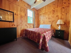 A bedroom with wooden paneling, a bed with a patterned bedspread, a nightstand with a lamp, and a ceiling fan. - Cozy Chalet #6/ Heated Pool/ Close To Beach/ On Site Activities! (Brackley Beach)