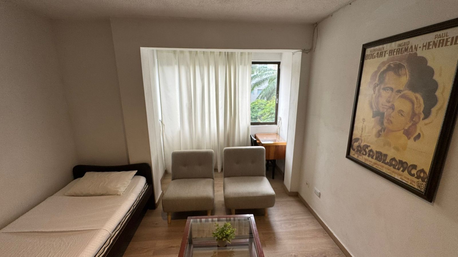 A small, furnished room with a bed, two chairs, a table, and a window with curtains.