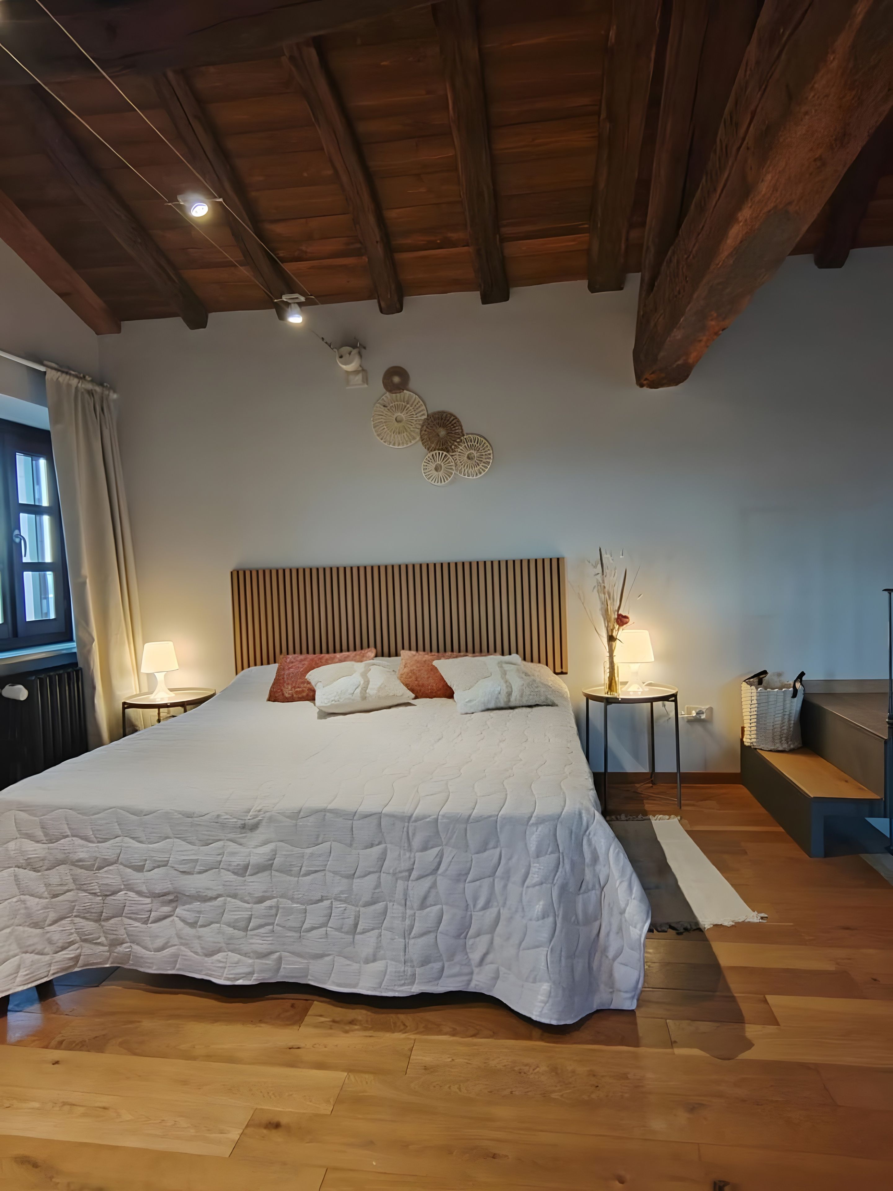 Un dormitorio con una cama grande, vigas de techo de madera, una mesita de noche con lámpara y una ventana con cortinas.