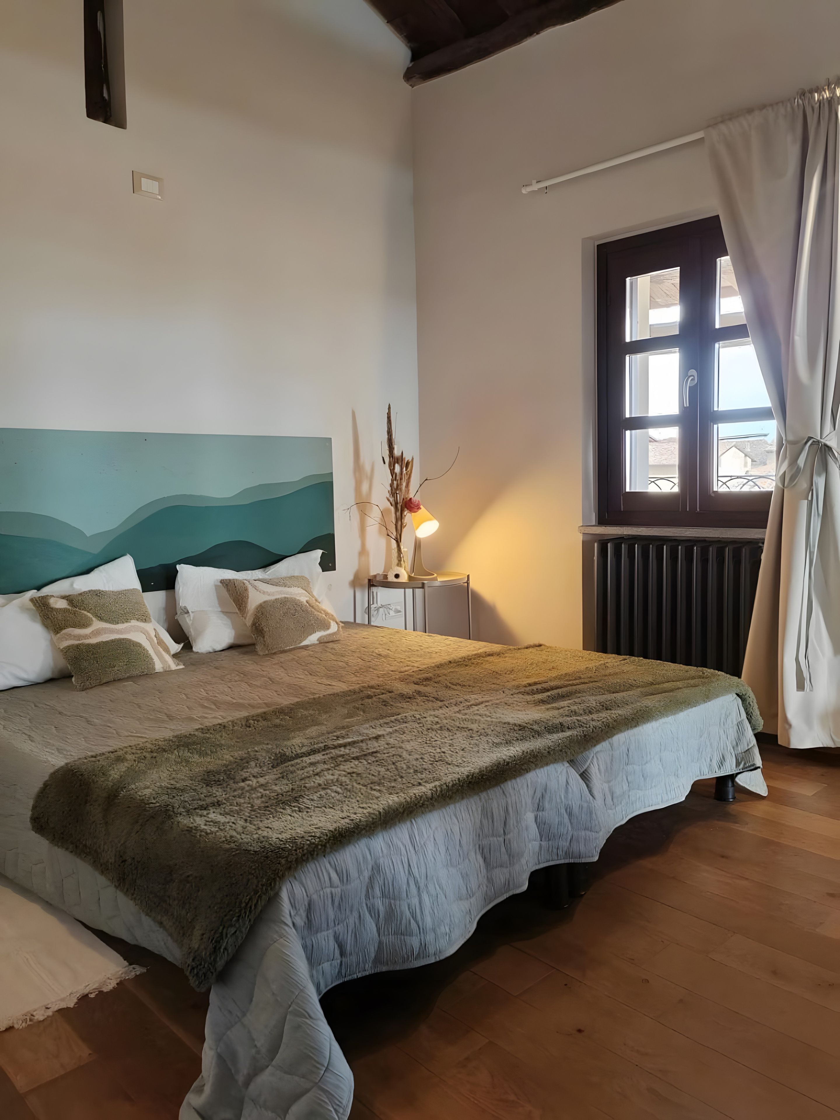 A bedroom with a bed, a nightstand, a window with curtains, and a wall with a painted design.