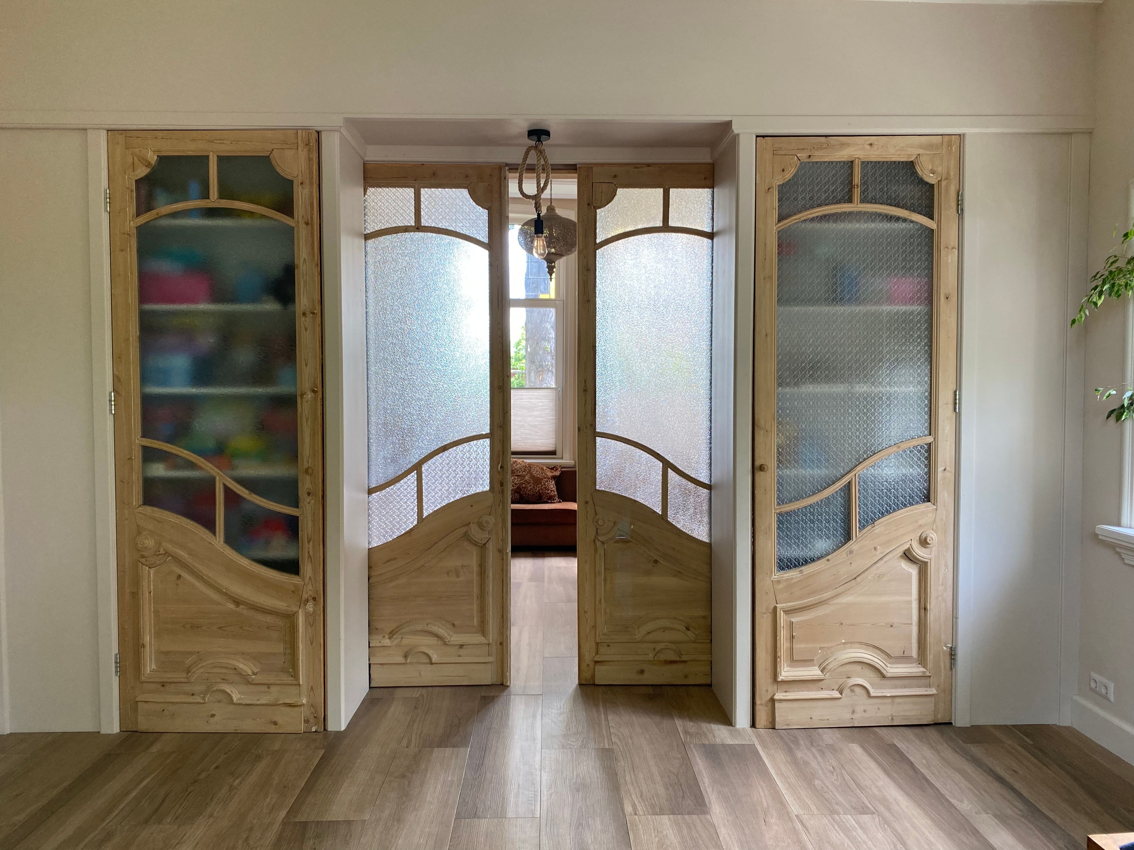 A hallway with wooden doors leading to a room, a staircase, and a window with a view.