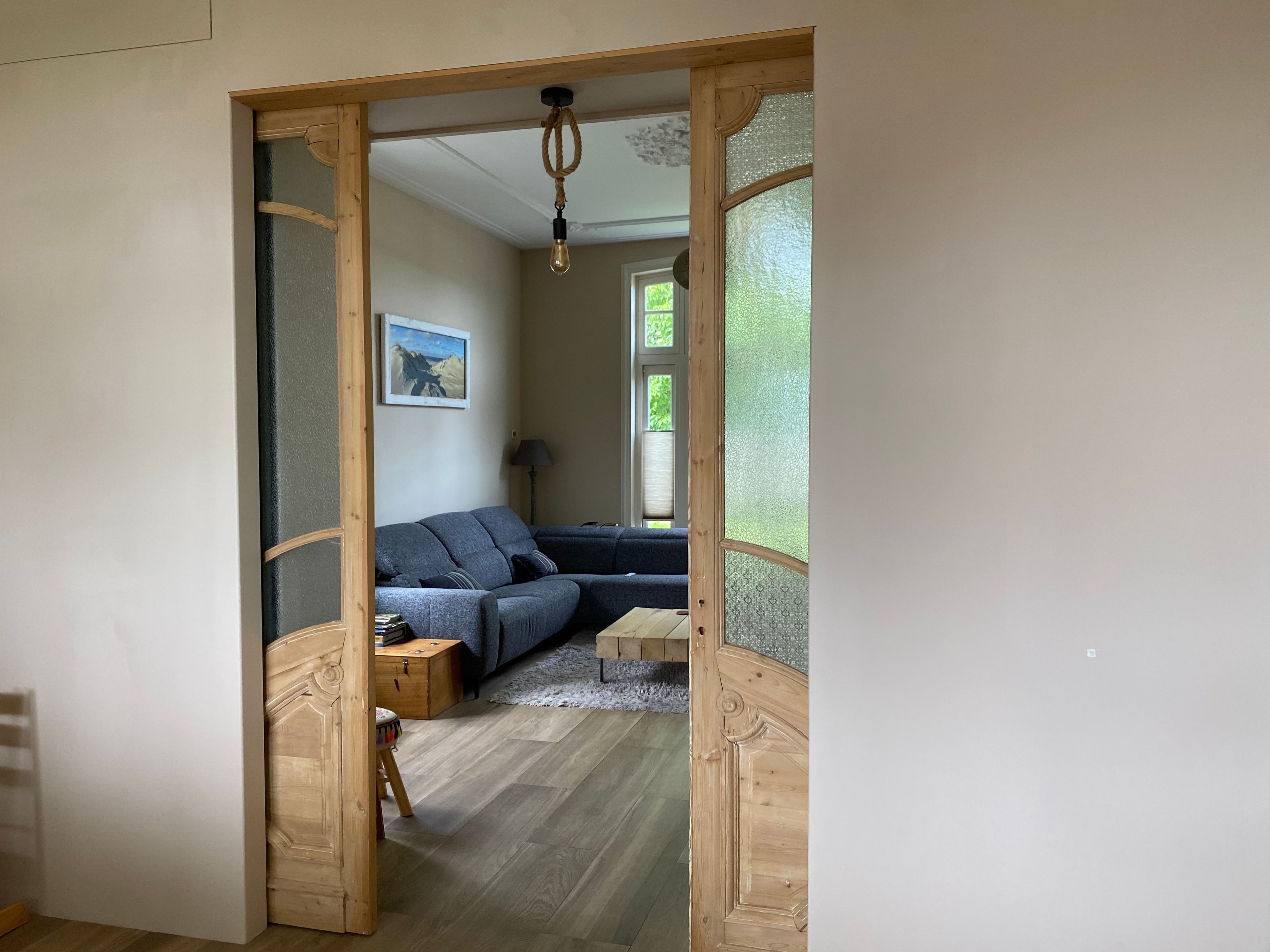 A room with a wooden door leading to a living area with a sofa, a lamp, and a painting on the wall.