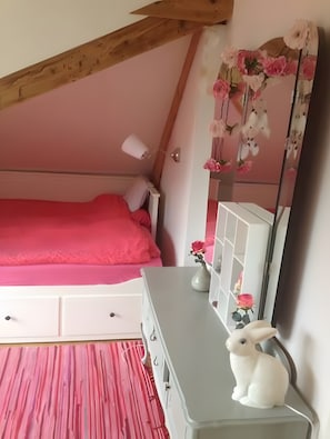 Un dormitori amb sostre inclinat, un llit amb estriat rosa, una petita còmoda amb mirall i una composició floral penjada a la paret.