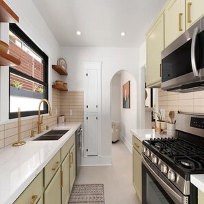 A modern kitchen with white cabinetry, stainless steel appliances, and a window with a view of greenery. - Spanish Retreat Two Houses Together (San Diego)
