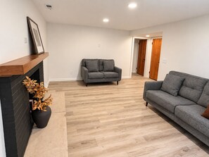 Een woonkamer met een open haard, twee grijze banken en een houten vloer.