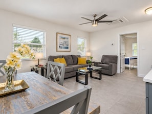 A modern living room with a gray sofa, a dining table with chairs, a ceiling fan, and a view of the outside through large windows. - Modern 1BR Steps to Madeira Beach with Courtyard and Grill (Madeira Beach)
