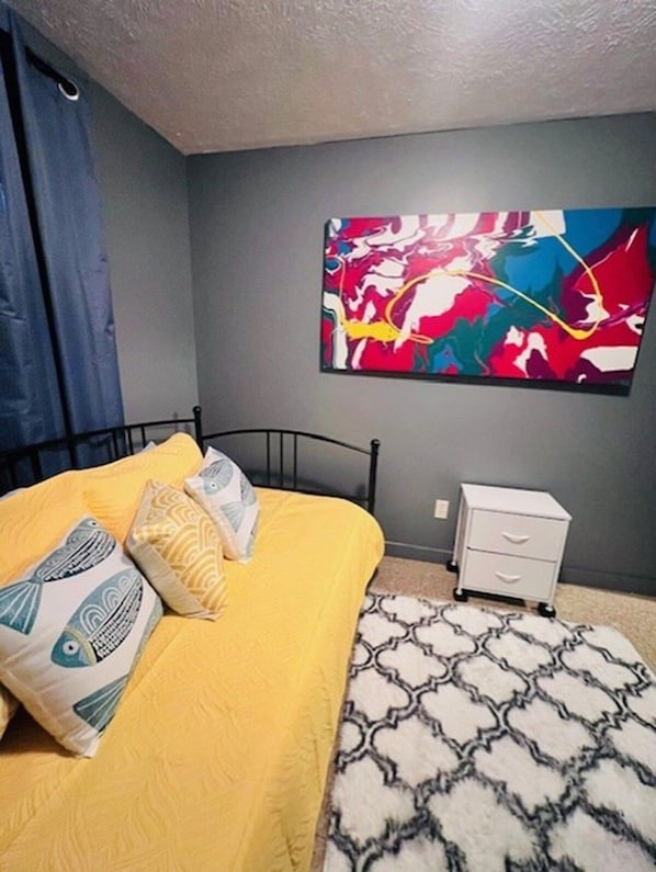 A bedroom with a bed, a nightstand, a colorful abstract painting, and patterned rugs.