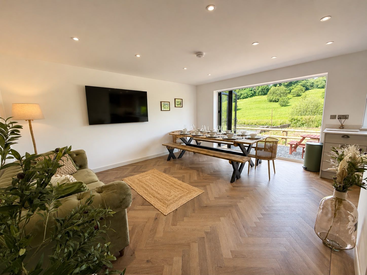 Ein modernes Wohnzimmer mit Fischgrätparkett, einer Couch, einem Fernseher und einem Essbereich mit Blick auf eine grüne Landschaft.