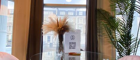 A room with a glass table, two pink chairs, a vase with dried plants, a QR code sign, and a view of buildings through a window.