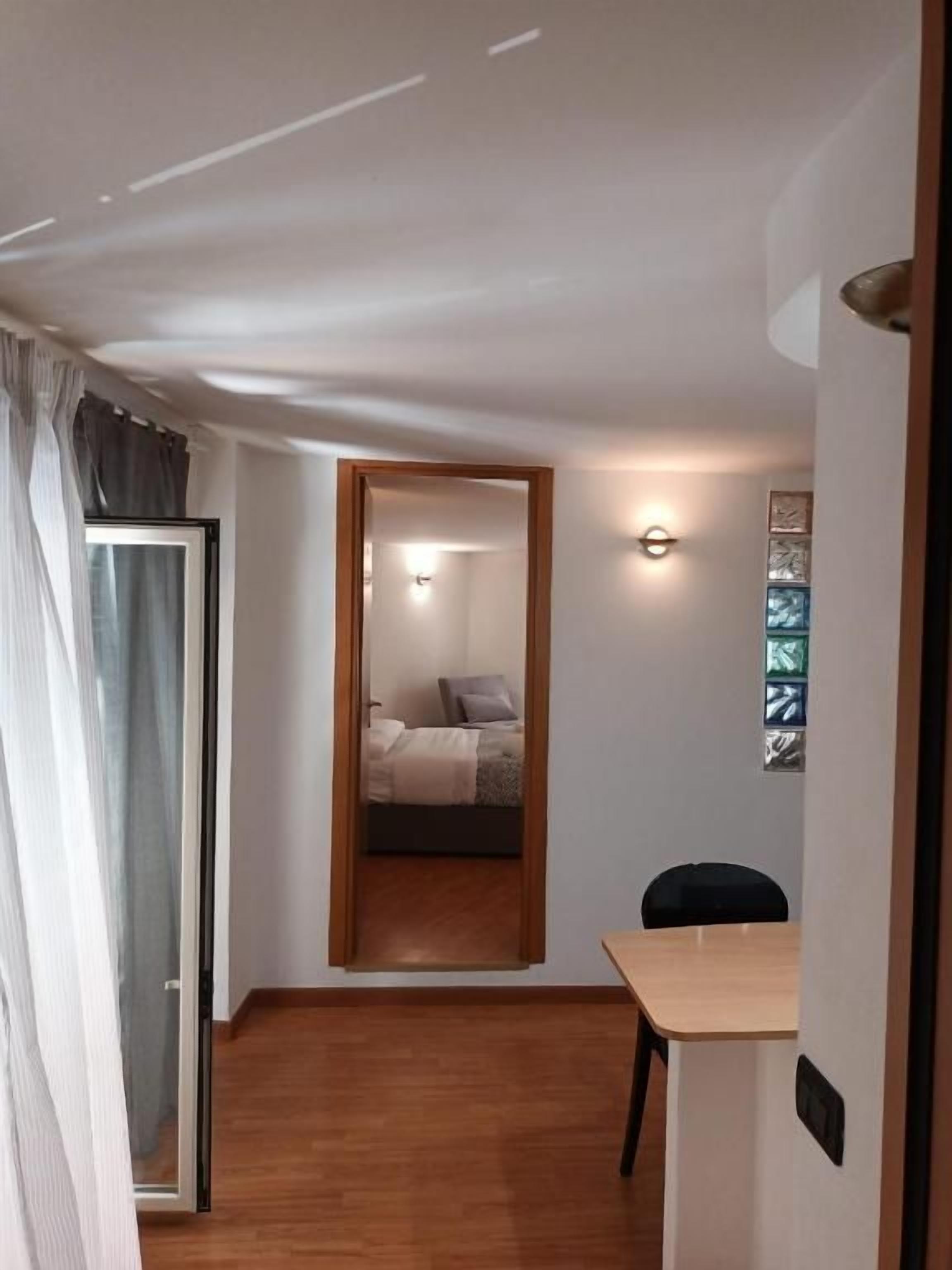 Un pasillo estrecho con piso de madera, una cama con cabecera blanca, una mesita con una silla y un aplique en la pared.