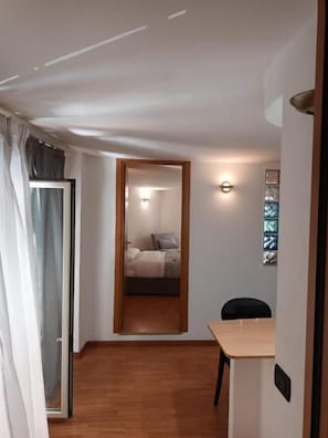 Un pasillo estrecho con piso de madera, una cama con cabecera blanca, una mesita con una silla y un aplique en la pared.