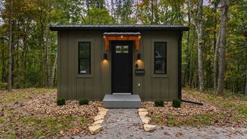 A small, single-story cabin with a black door and two windows, surrounded by a forest.