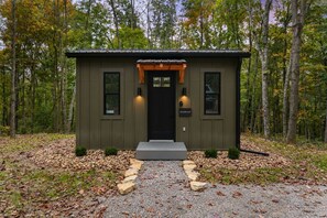 Eine kleine, eingeschossige Holzhütte mit einer schwarzen Tür und zwei Fenstern, umgeben von einem Wald.