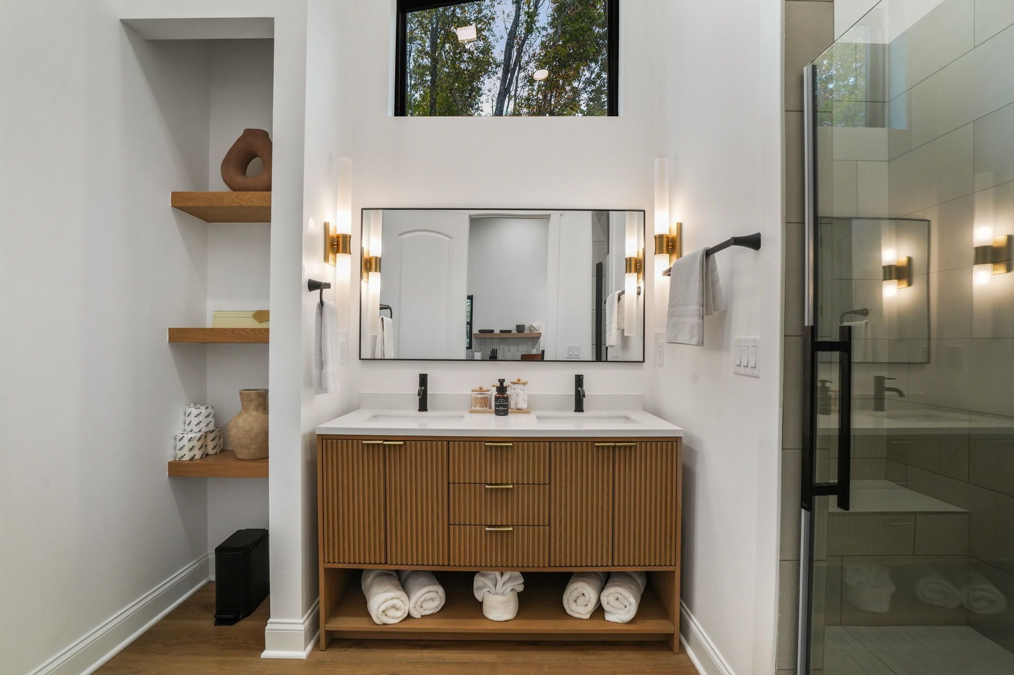 A modern bathroom with a large mirror, wooden vanity, and built-in shelves.