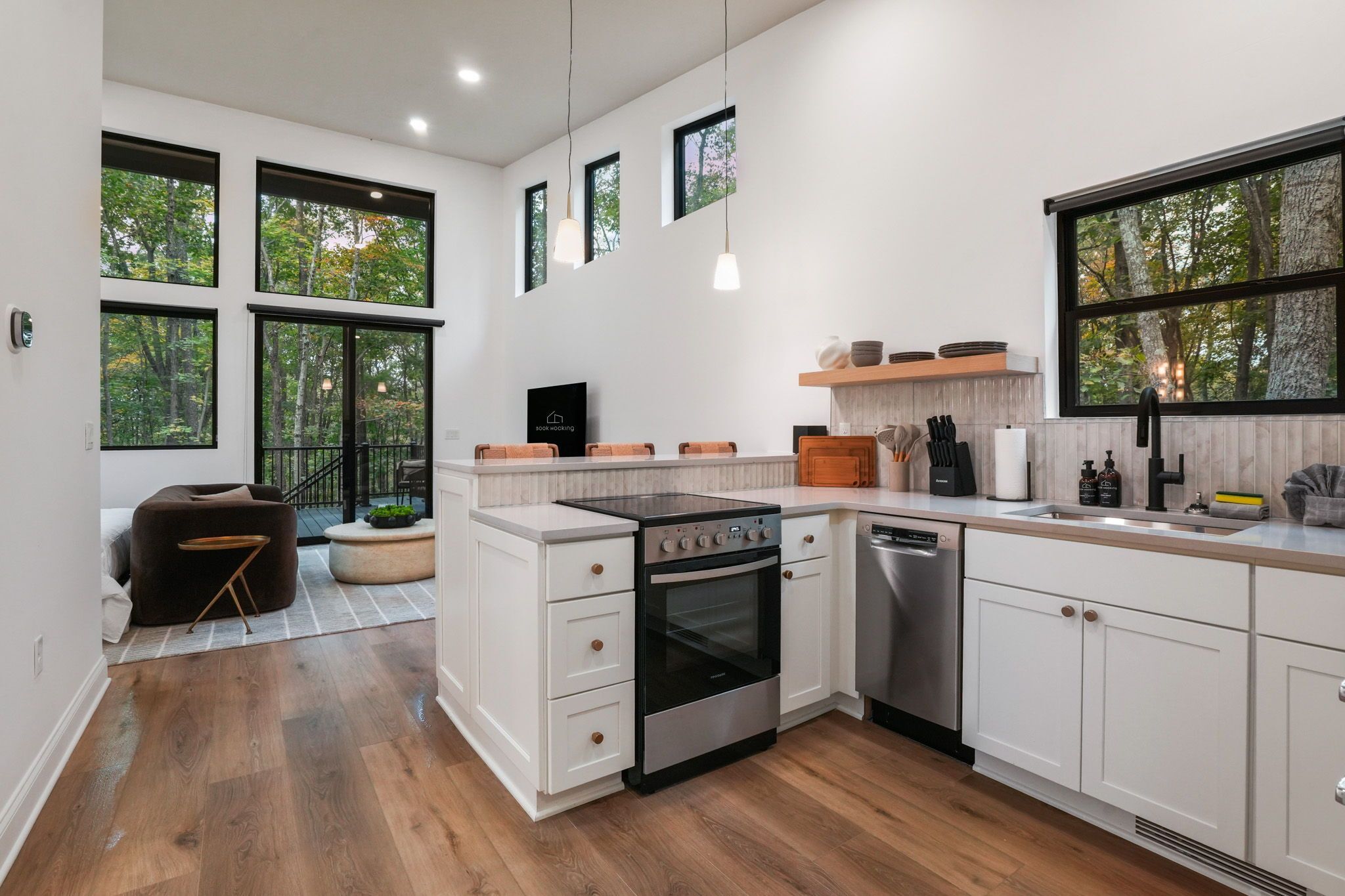 A modern kitchen with white cabinetry, a built-in oven, and a dishwasher. There are large windows providing natural light and views of trees.