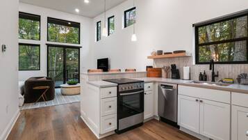A modern kitchen with white cabinetry, a built-in oven, and a dishwasher. There are large windows providing natural light and views of trees.
