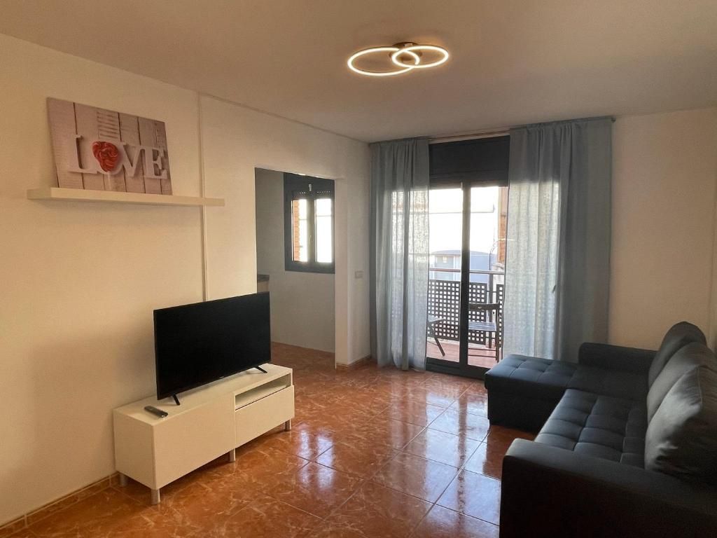 Una sala de estar con un televisor de pantalla plana sobre un soporte blanco, un sofá oscuro, una puerta corrediza de vidrio que da a un área exterior, y una lámpara de pared.