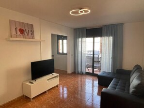 Una sala de estar con un televisor de pantalla plana sobre un soporte blanco, un sofá oscuro, una puerta corrediza de vidrio que da a un área exterior, y una lámpara de pared.