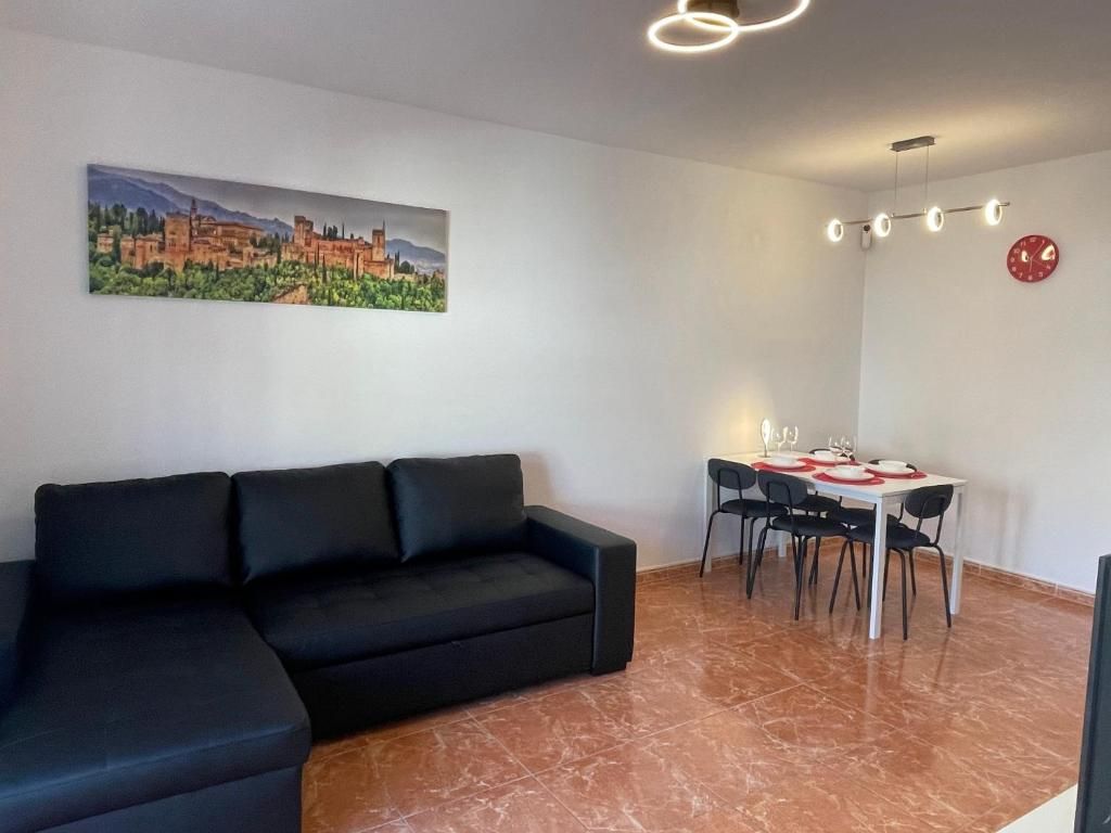 Una sala de estar con un sofá negro, una mesa de comedor con sillas, un cuadro en la pared y un reloj.