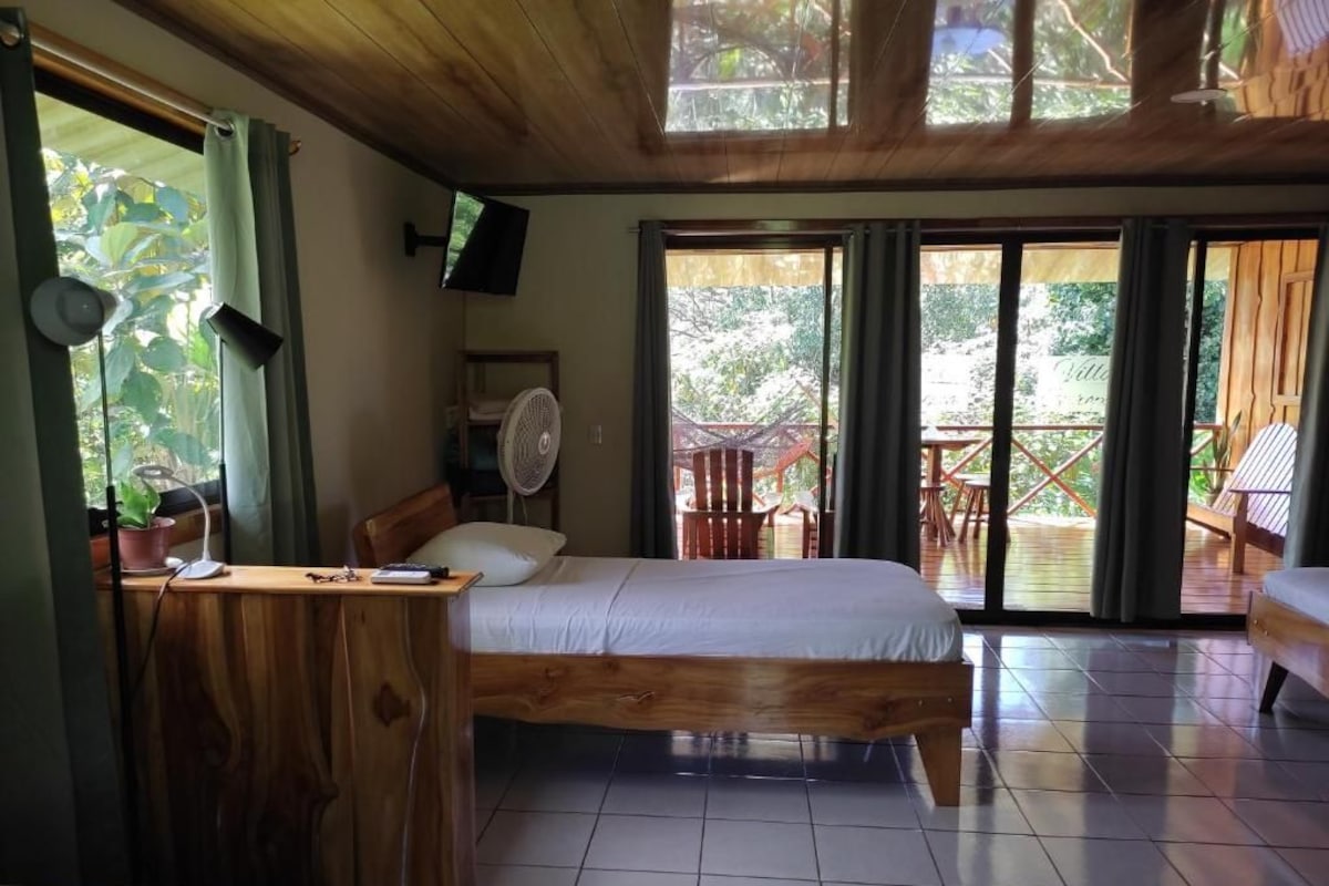 A room with a bed, a desk with a chair, a fan, and a television. There are sliding glass doors leading to an outdoor area with chairs and a table.