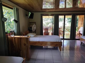 A room with a bed, a desk with a chair, a fan, and a television. There are sliding glass doors leading to an outdoor area with chairs and a table. - Cecropia Wild Corcovado (Drake)