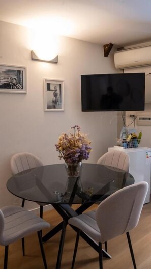 Ein Essbereich mit einem runden Glastisch, vier Stühlen, einer Vase mit Blumen, einem flachbildfernseher an der Wand und einem kleinen Kühlschrank.