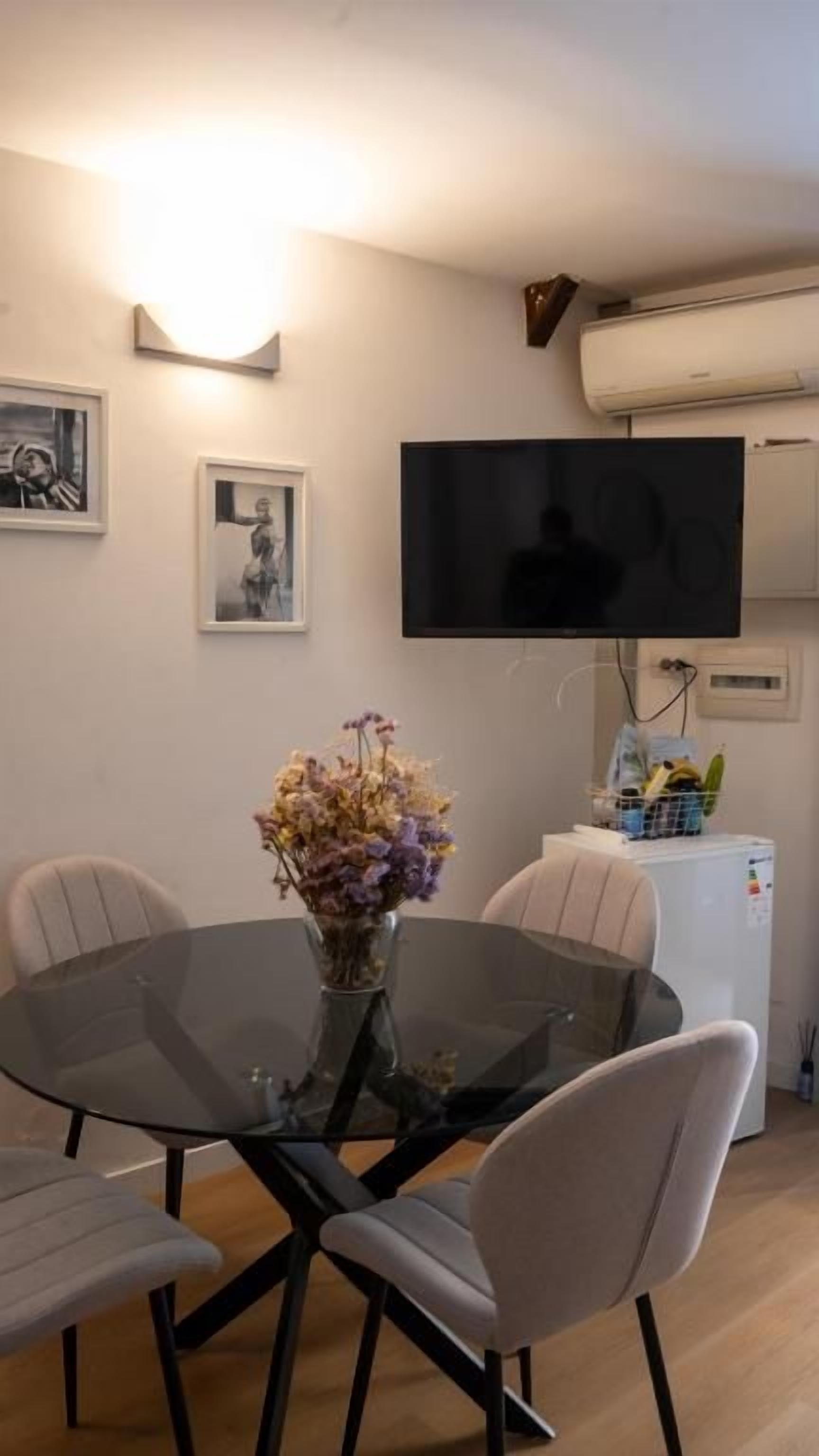 A dining area with a glass table, four chairs, a vase with flowers, a flat-screen TV mounted on the wall, and a white refrigerator.