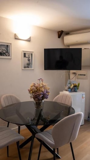 Uma sala de jantar com mesa de vidro, quatro cadeiras, um vaso com flores, TV de tela plana na parede e uma geladeira branca.
