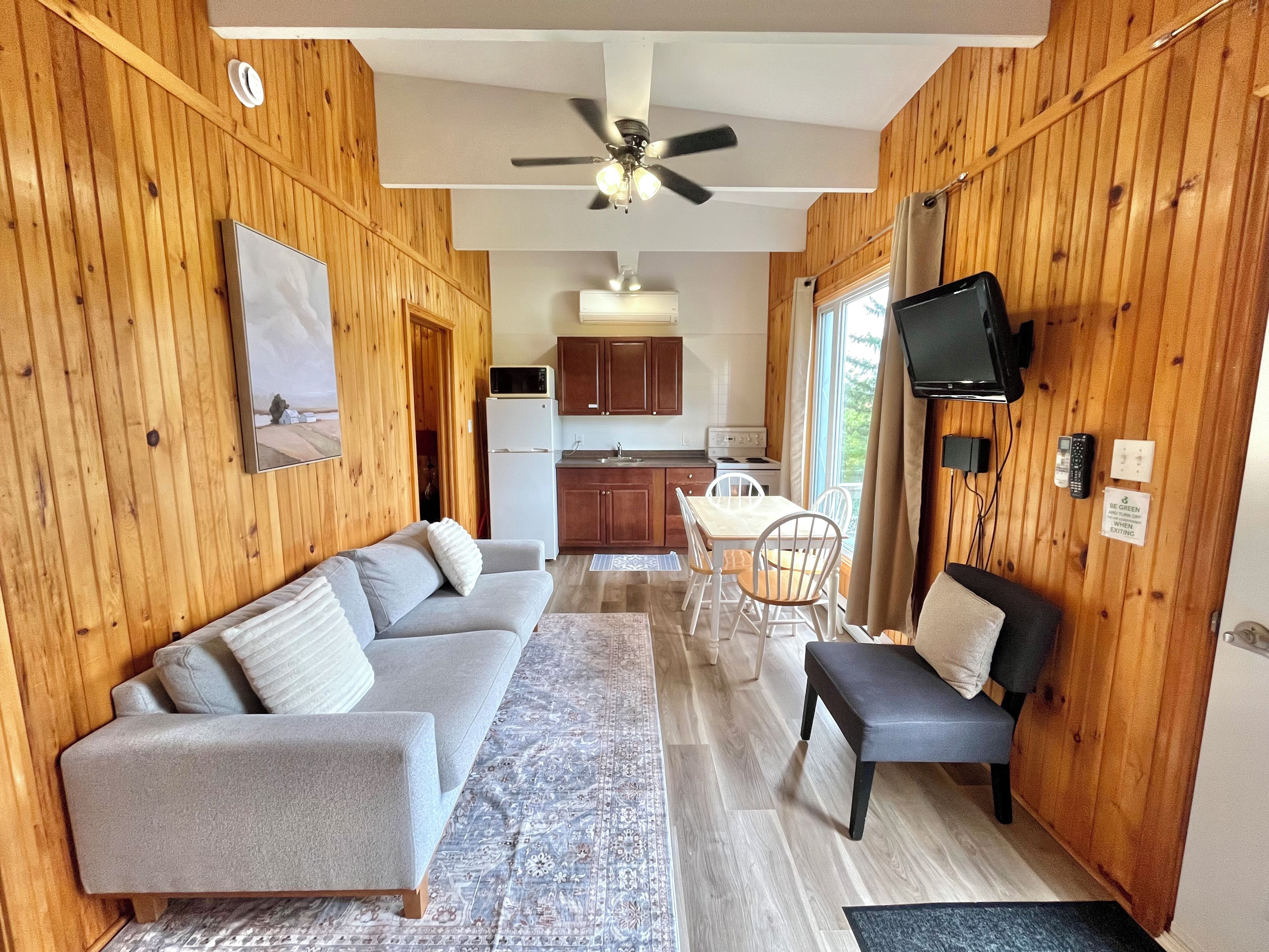 A compact living space with a sofa, a dining table with chairs, a television mounted on the wall, and a kitchen area with wooden walls and a ceiling fan.