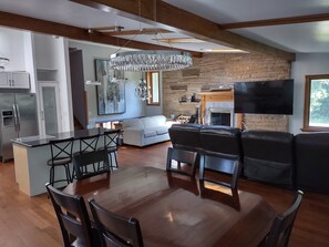 A modern kitchen with a built-in island, a dining area with a wooden table and chairs, a living room with a fireplace, and a large TV mounted on the wall. - Beautiful 6 Bedroom 3.5 Bath Home with Heated Pool and Jacuzzi and Steam Rooms (Hales Corners)