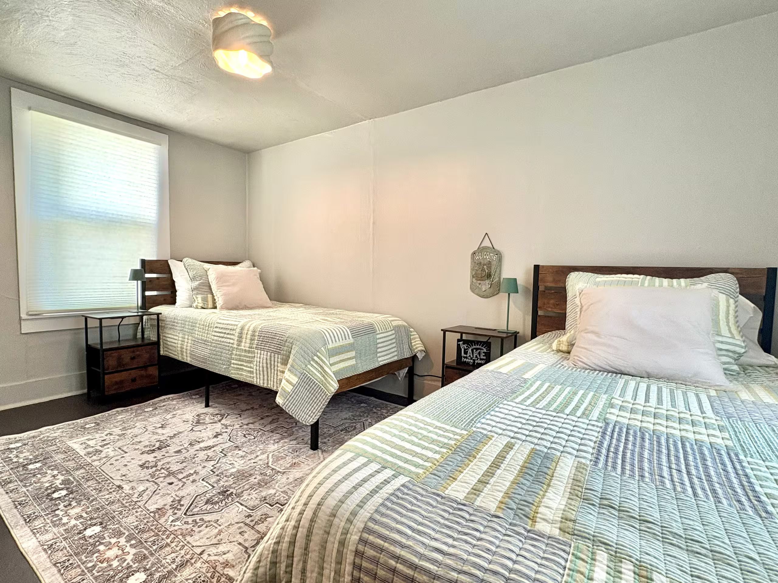 A bedroom with two beds, a patterned rug, a wall-mounted light fixture, and a window with blinds.