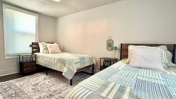 A bedroom with two beds, a patterned rug, a wall-mounted light fixture, and a window with blinds.