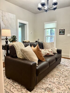 A living room with a dark brown leather sofa, patterned rug, and a chandelier. - The Drake on Main Street - Charming & Convenient - Downtown Home by the Harbor (Warsaw)