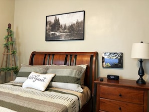 Una cama bien hecha con una almohada que dice 'Our Guest', un cuadro de paisaje enmarcado encima, una mesita de noche con una lámpara y una escalera decorada con follaje contra la pared.