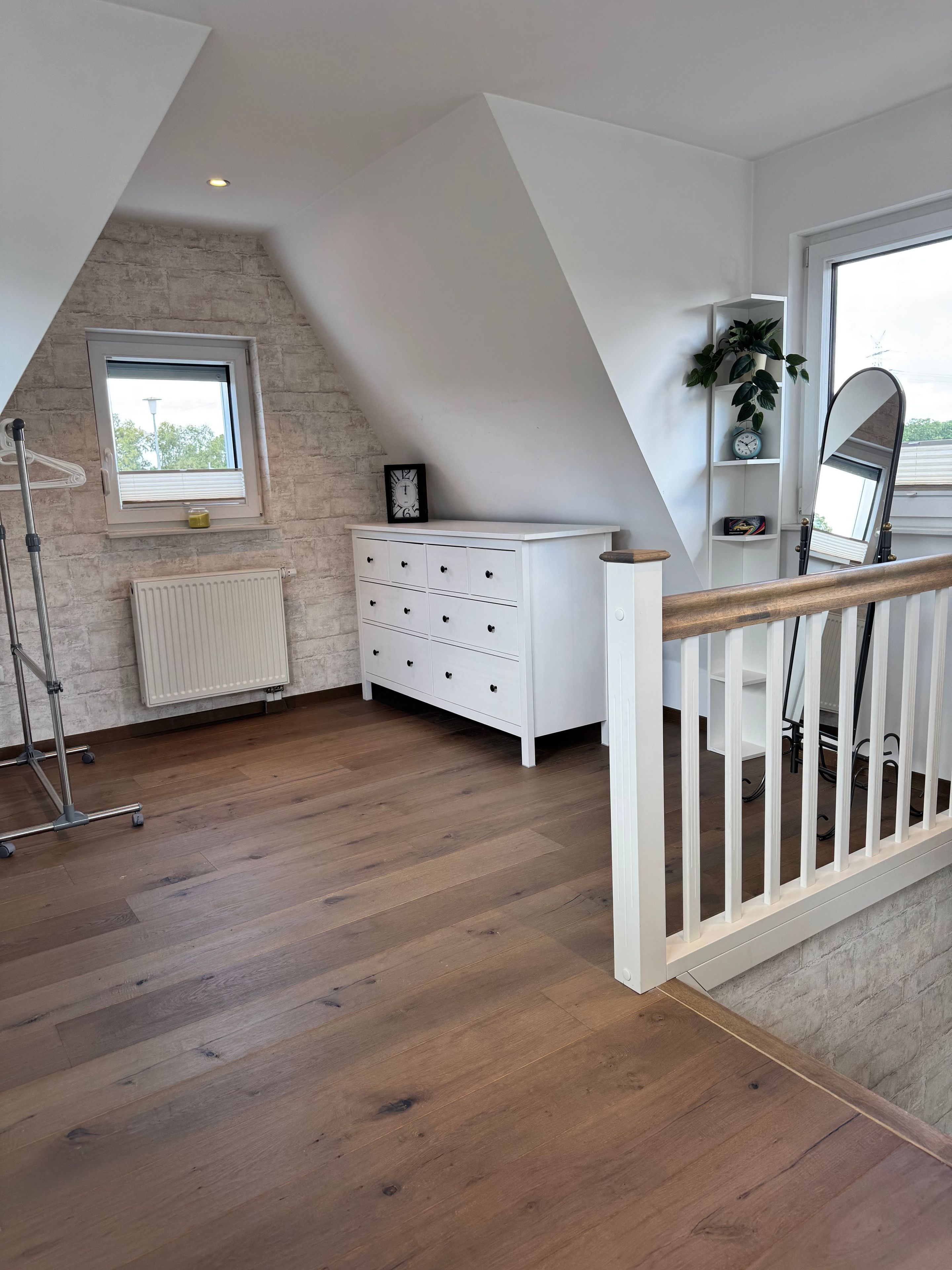 A room with a sloped ceiling, a white dresser, a radiator, a wooden floor, and a window with a view.