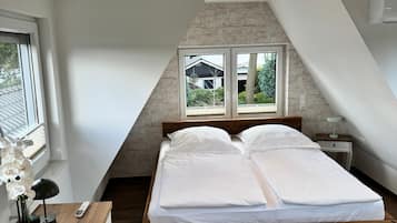 A bedroom with a sloped ceiling, a bed with white bedding, a nightstand, a lamp, and a window with a view of a house and trees.