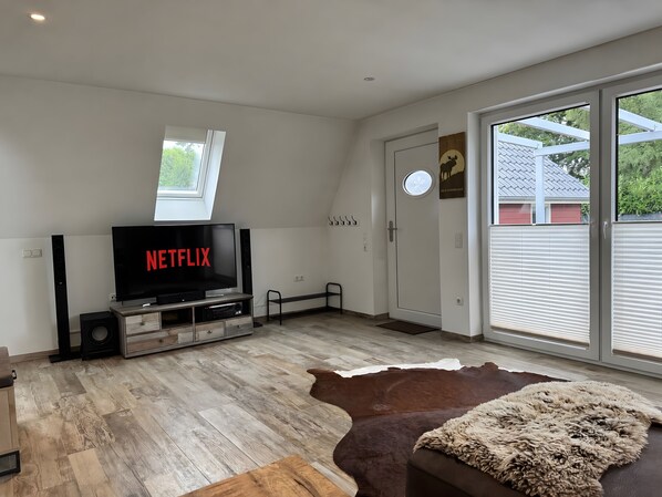 En moderne stue med et fladskærms-tv der viser 'NETFLIX', et træunderholdningsmøbel, et stort vindue og en kohude-gulvtæppe.