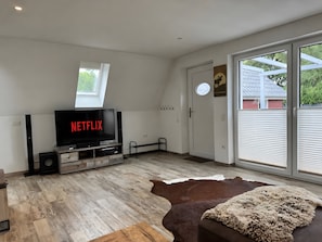 Un salon moderne doté d’un téléviseur à écran plat affichant le logo « NETFLIX », d’un meuble de rangement en bois, d’une grande fenêtre et d’un tapis en peau de vache.