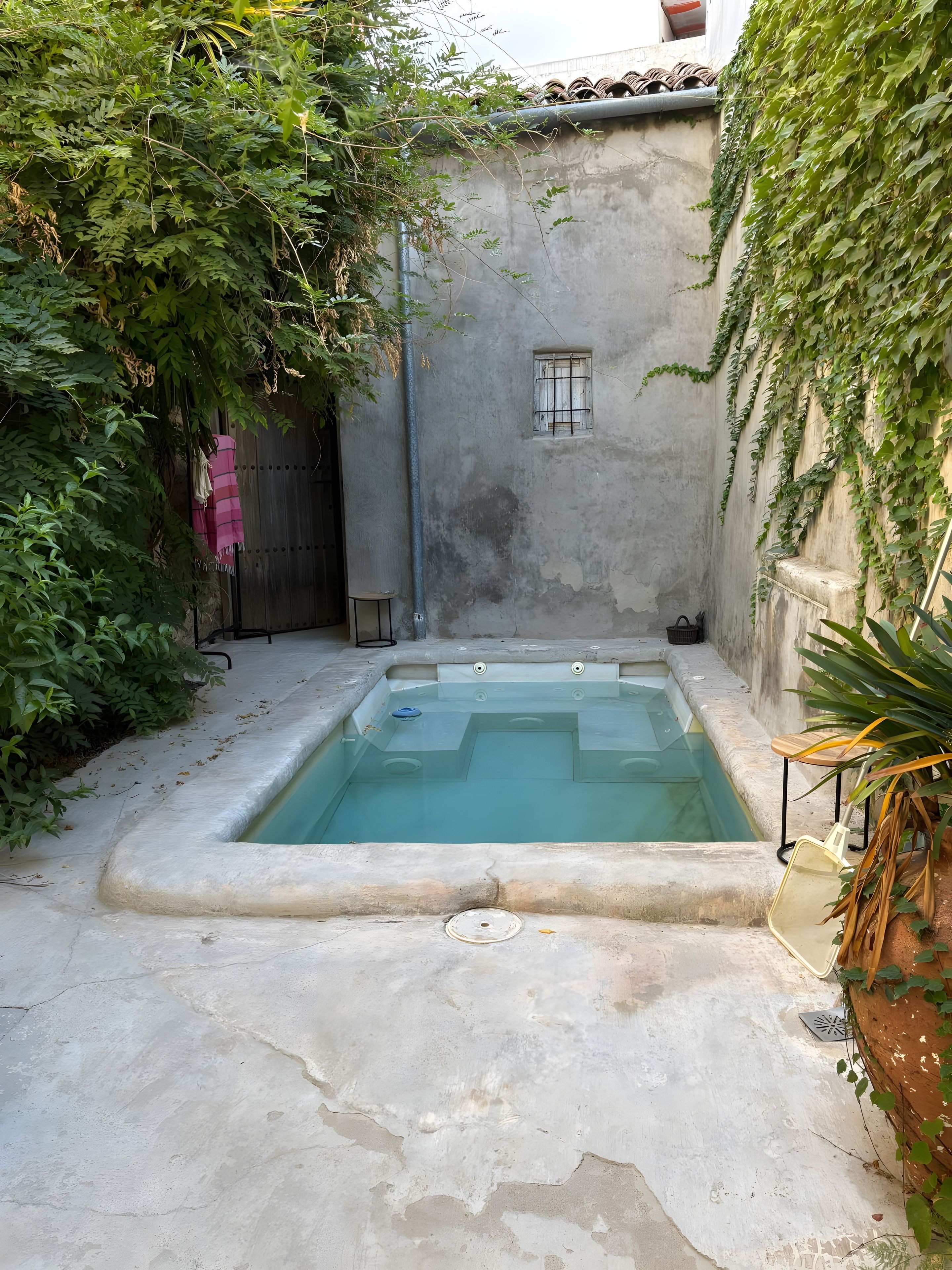 Una piscina rectangular con borde de hormigón, rodeada de suelo de hormigón, un ventanal y una puerta con un paño rosa.