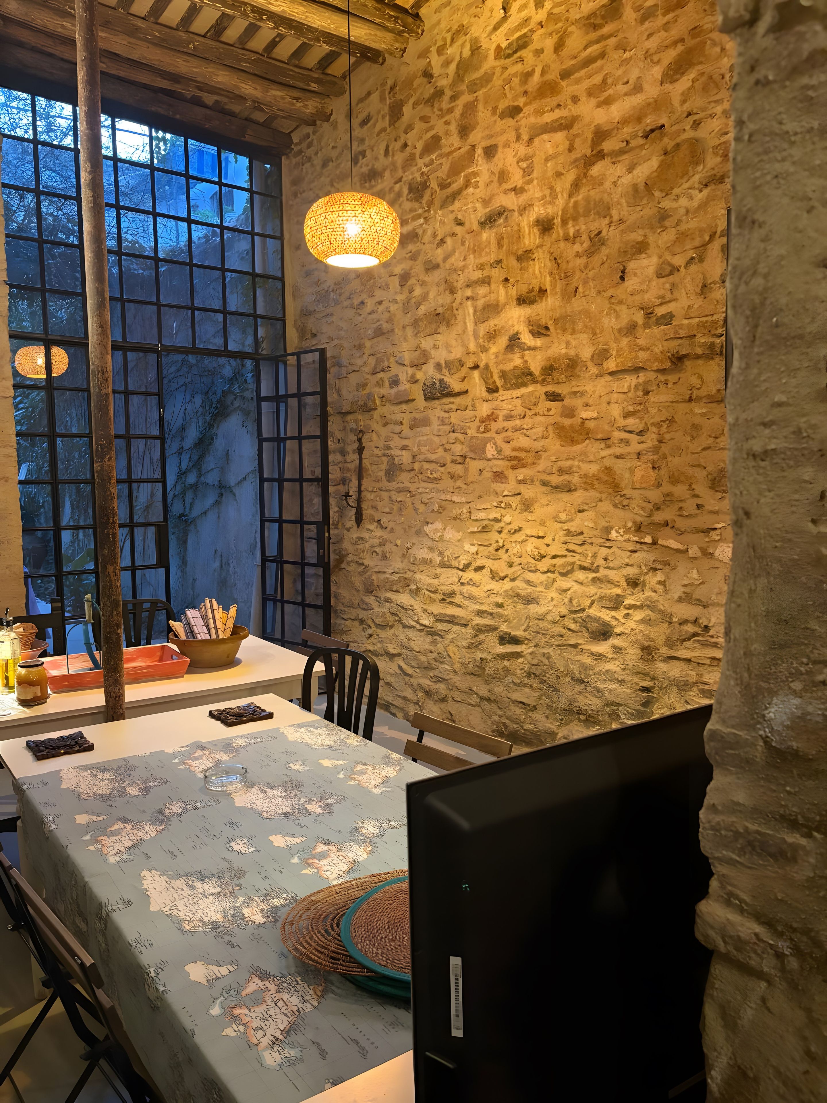 Un comedor rústico con pared de piedra, un ventanal grande y una mesa dispuesta con pan, un bol y una botella.