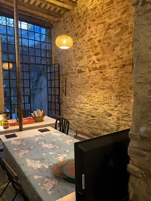 Un comedor rústico con pared de piedra, un ventanal grande y una mesa dispuesta con pan, un bol y una botella.