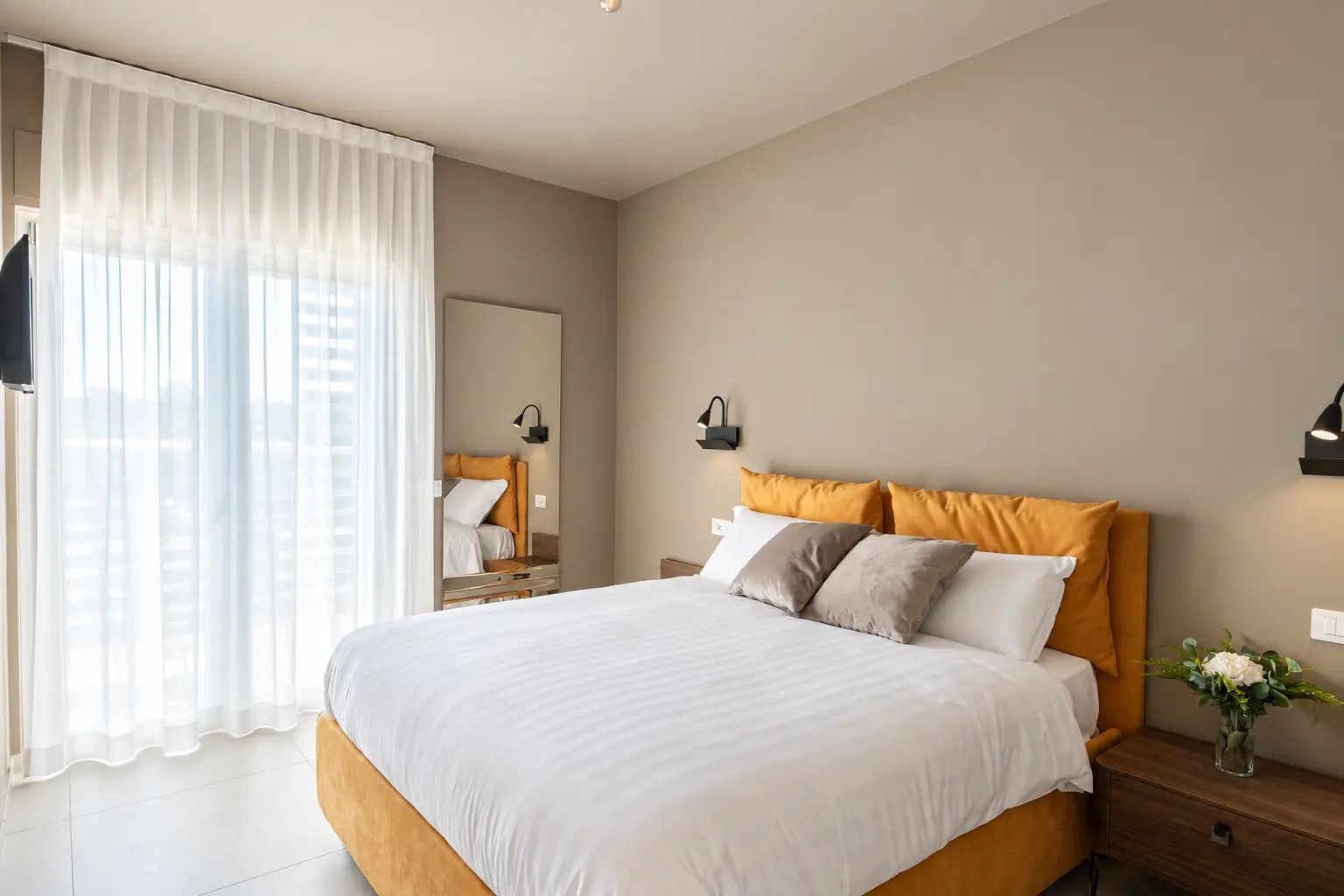 A modern bedroom with a large bed, a nightstand, a mirror, and a window with sheer curtains.