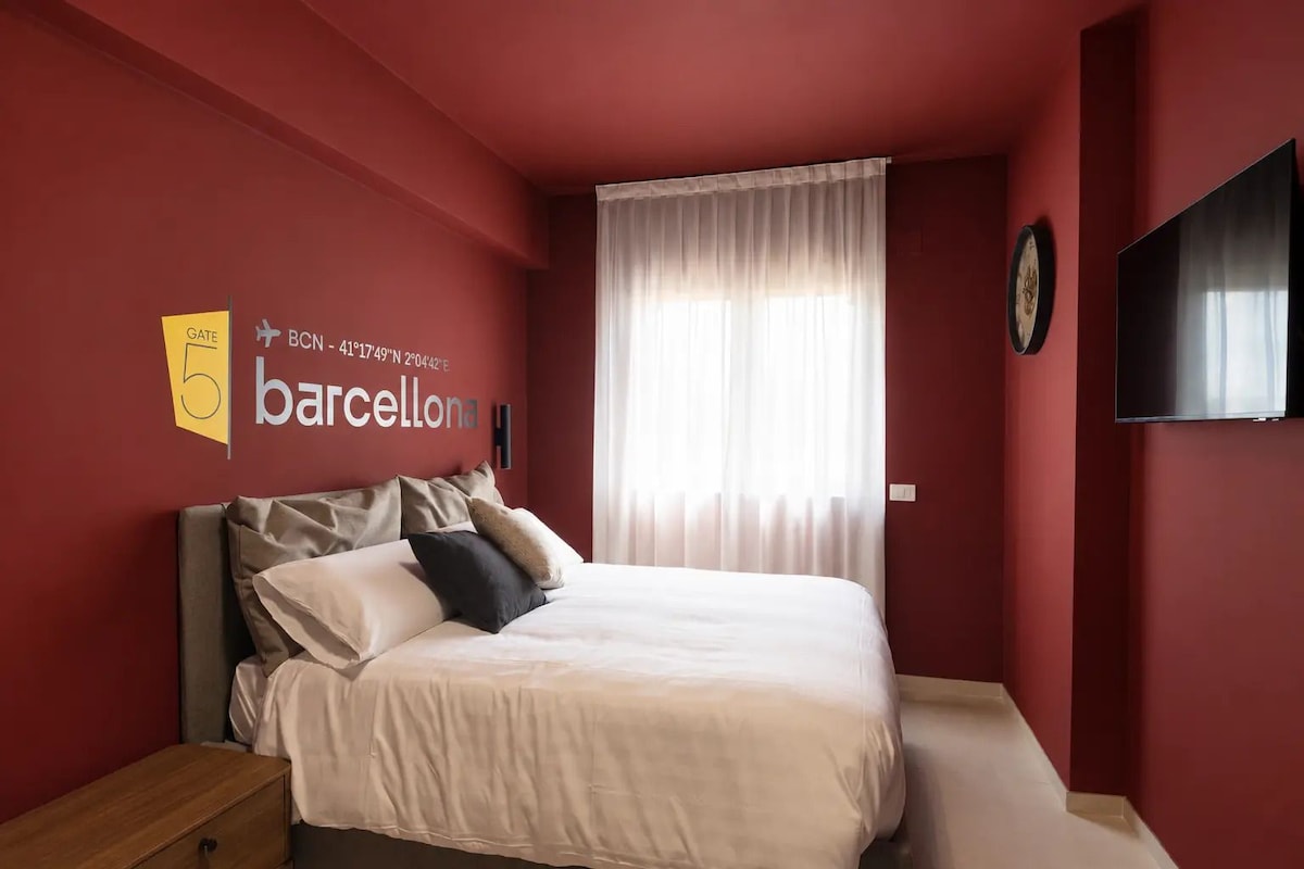 Habitació d'hotel amb un llit, una tauleta de nit, un rellotge de paret i una finestra gran amb cortines.