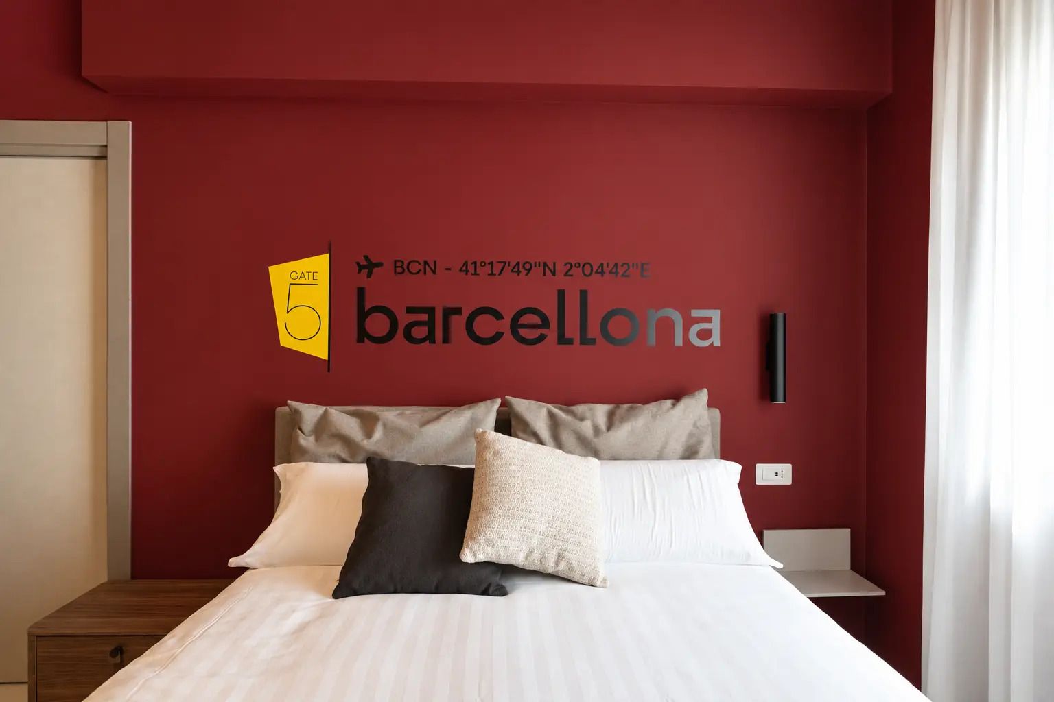 Habitación de hotel con pared roja, cama con ropa blanca y señalización que indica la ubicación como BCN - 41°17'49'N 2°04'42'E.