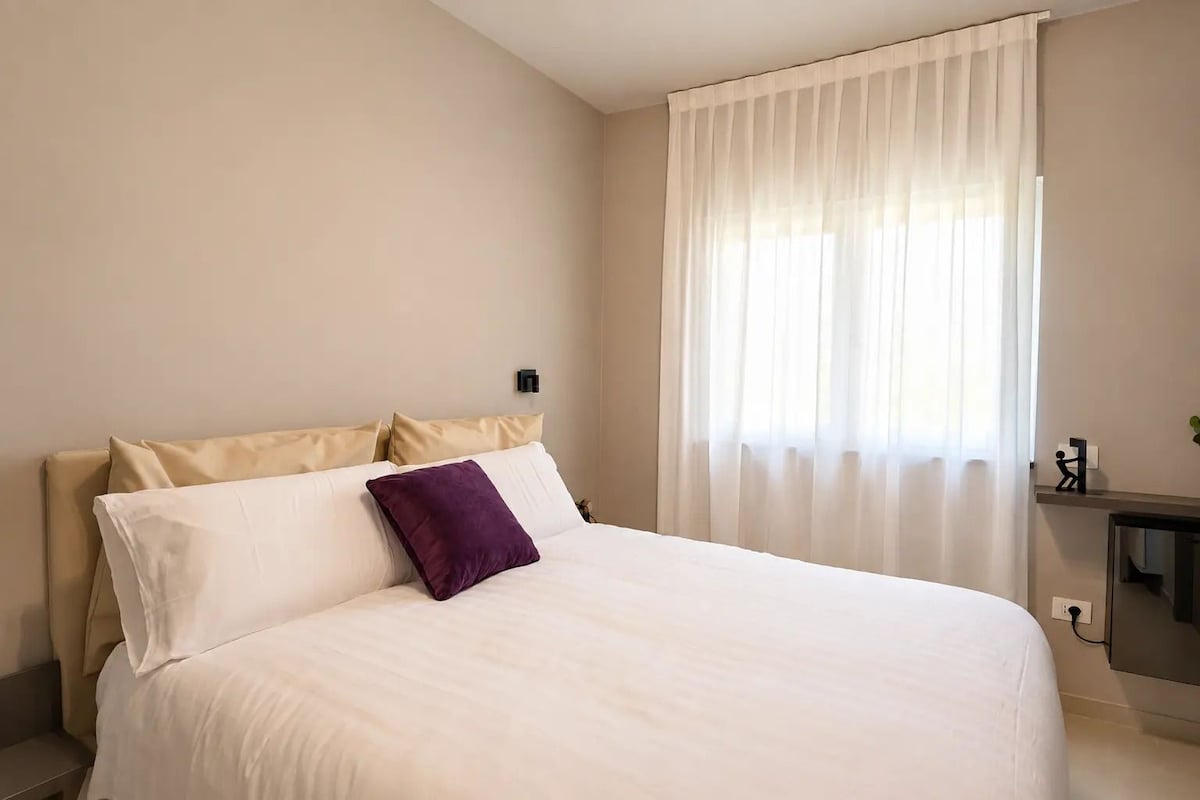 Habitació d'hotel amb llit, una vetust i una finestra amb cortines translúcides.