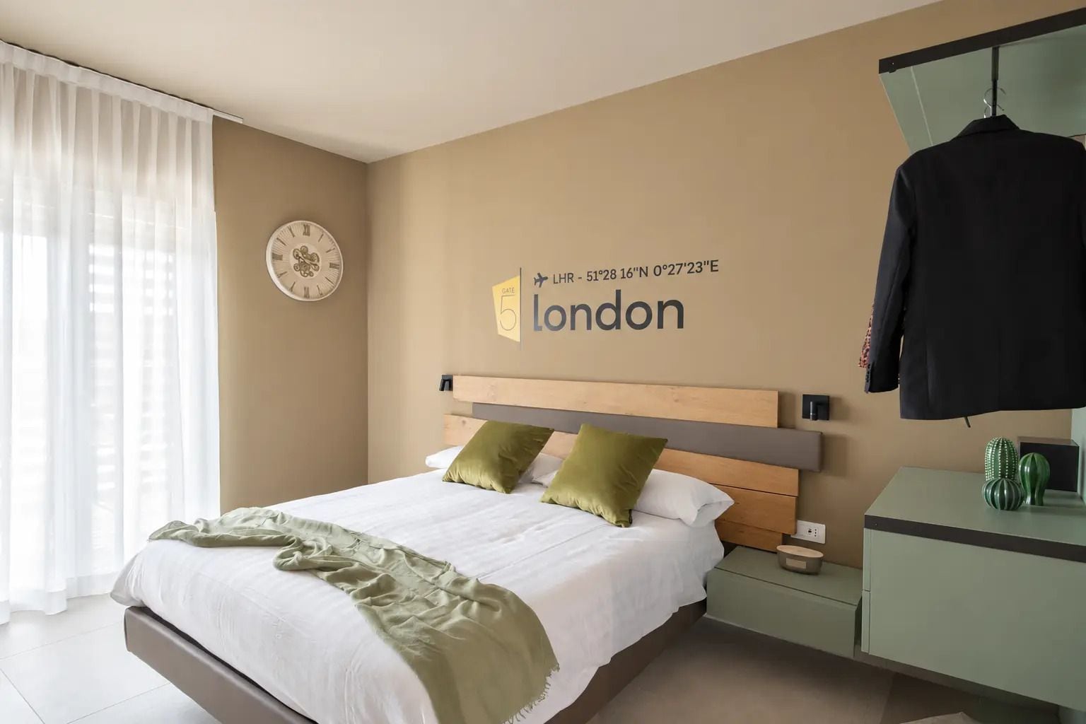 Una habitación de hotel con una cama, un reloj, un cartel que indica Londres como ubicación, y un abrigo colgado en la pared.