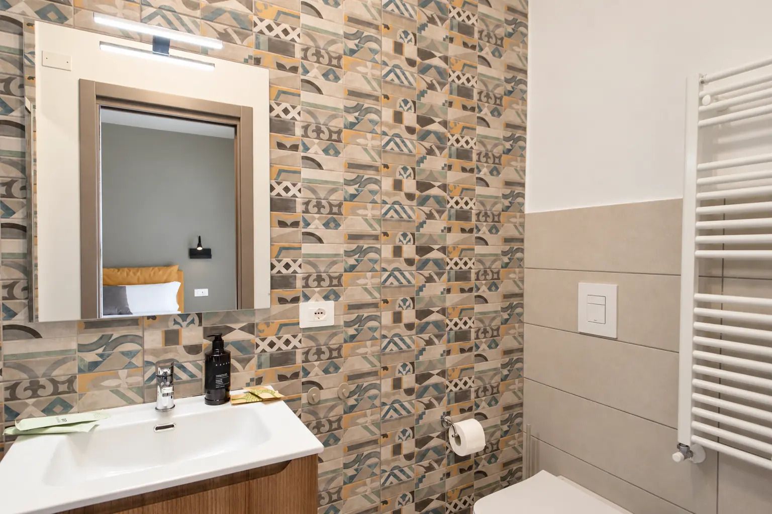 Une salle de bain avec un mur recouvert de carreaux décoratifs, un miroir au-dessus du lavabo, un distributeur de serviettes et des toilettes avec un porte-serviettes.