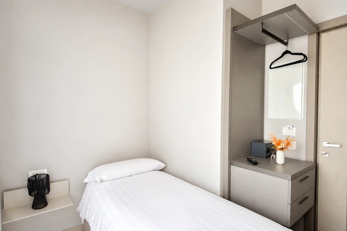 Habitació d'hotel minimalista amb un llit individual, una taula de nit amb comandament a distància, un vas petit amb flors i una perxa penjant.